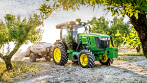 Zeleno rumen traktor John Deere 5090EN na sončen dan vleče pršilnik skozi sadovnjak