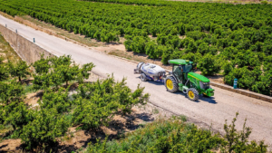 Zelen traktor John Deere 5090EN, ki vleče pršilnik po kmetijski poti v bližini sadovnjakov