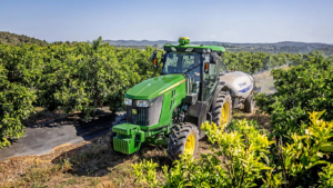 Zeleno rumeni traktor John Deere 5105EN vleče pršilnik skozi sadovnjak z bujno rastjo, za njim se razprostirajo valoviti griči