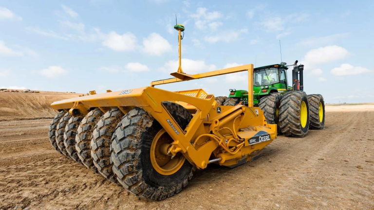 Traktor John Deere med vleko velikega rumenega priključka za oranje z več težkimi kolesi pri obdelavi suhih tal