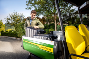 Oseba, ki naklada velik črn kovček na keson vozila John Deere Gator GS s svetlo rumenimi sedeži, obkroženim z zelenjem in drevesi.