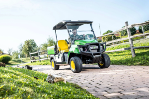 Zeleno večnamensko vozilo John Deere Gator GS vozi po tlakovani poti po urejeni zelenici z lesenimi ograjami.