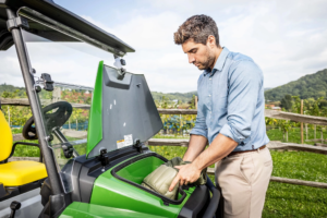 Oseba, ki odlaga oblazinjen nahrbtnik v sprednji predal zelenega vozila John Deere Gator GS, parkiranega na prostem ob leseni ograji in vinogradu.