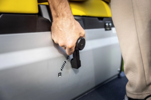 Bližnji posnetek roke, ki upravlja prestavno ročico vozila John Deere Gator GS z označenima položajema za vožnjo naprej in nazaj ob rumenih sedežih.