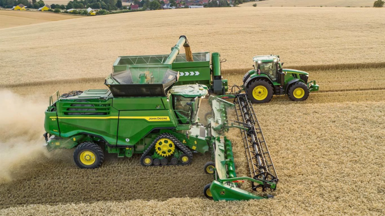 Kombajn John Deere med žetvijo žita in sočasnim razkladanjem v traktorsko prikolico, pogled od zgoraj na velikem polju